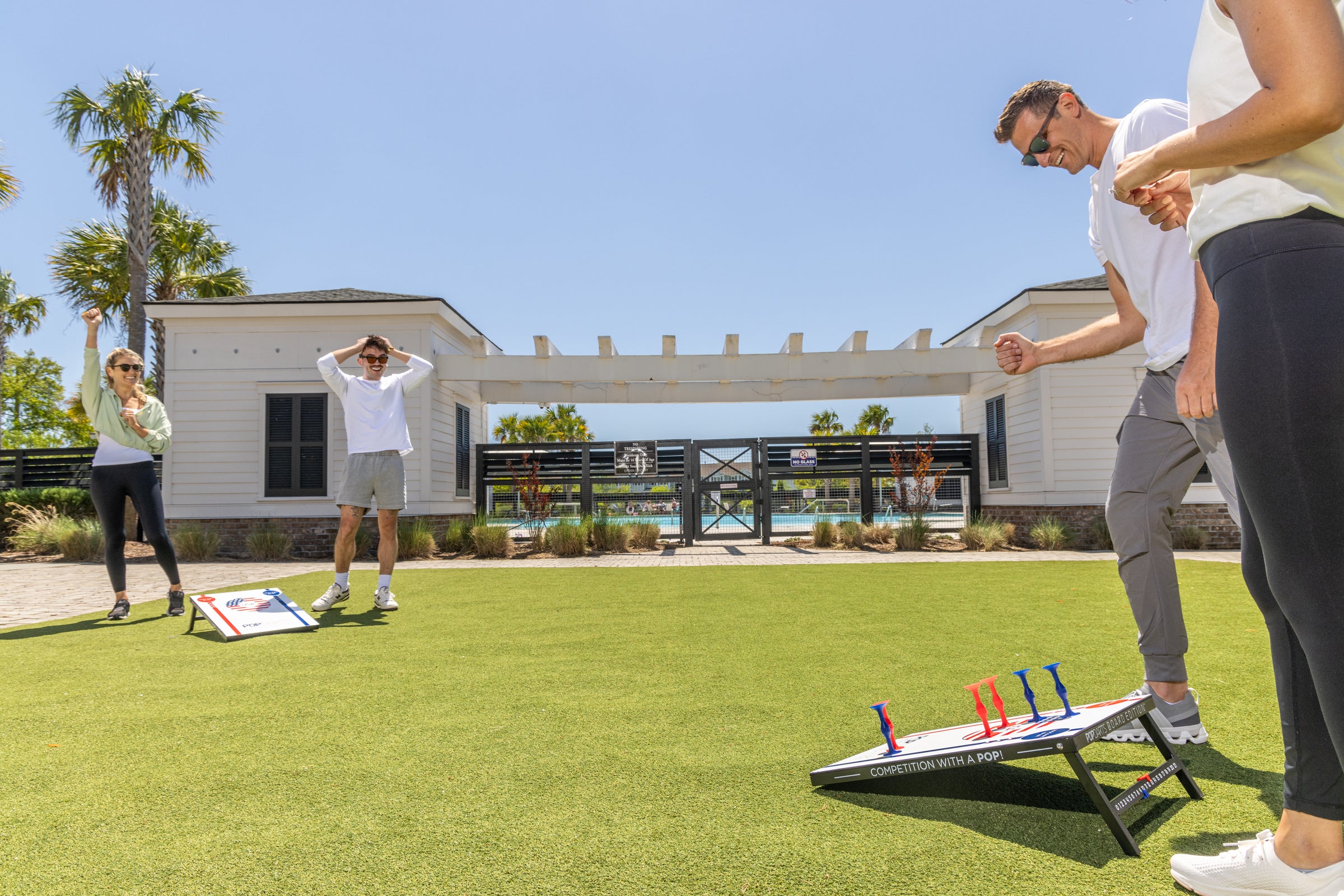 Real lifestyle photo: friends playing cornhole-style Popdarts board edition on a lawn next to a pool house, mid-celebration.